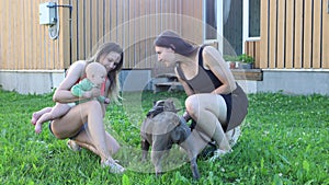 Two white women with an infant and dog socialising on grass outside rural house.