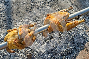 Two whole chickens roasted and grilled on a spit broach over a fire, ember and ashes