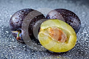 Two whole and one half sliced Damson plums with the stone on a black cutting board with water drops. Macro shot of fresh, organic