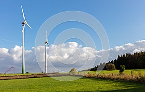 Two wind turbines in front of a band of clouds in a rural area on the edge of a forest.