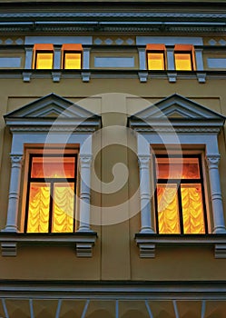 Two windows framed by facade decor, curtained with curtains with lambrequins shine yellow in the night