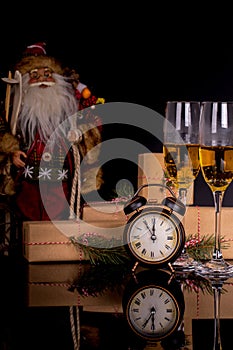 Two wine glasses with champagne, Santa Claus, clock and christmas gifts on a black background with reflection
