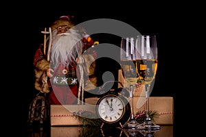 Two wine glasses with champagne, Santa Claus, clock and christmas gifts on a black background