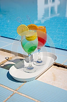 Two wine glasses with red cocktail or juice with bendie and piece of orange on edge, water and swimming pool behind