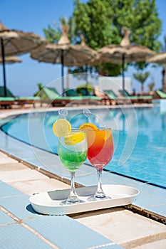 Two wine glasses with red cocktail or juice with bendie and piece of orange on edge, water and swimming pool behind
