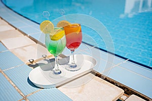 Two wine glasses with red cocktail or juice with bendie and piece of orange on edge, water and swimming pool behind