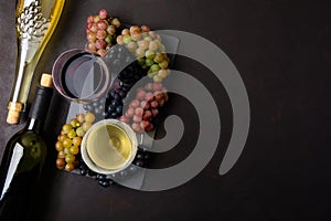 Two wineglasses with red and white wine, bottles and grapes on dark wooden background.