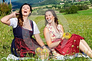 Two woman in dirndl sitting on blanket in meadow and laughing