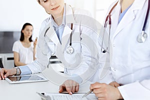 Two woman-doctors at work with patient at background. Female physicians filling up medical documents or prescription