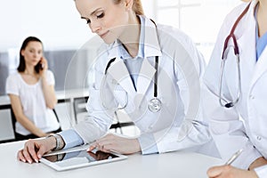 Two woman-doctors at work with patient at background. Female physicians filling up medical documents or prescription