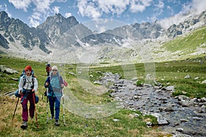Two woman hikers are walking with trakking poles by the river among mountains. Extreme tourists in wild nature