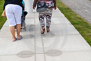 two Woman one with colorful trousers and wedge heel and one whit flipflop and white shorts pushes stroller, floor