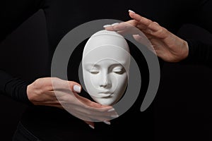 Two woman`s hands hold white gypsum mask face on a black background. Concept social psychological masks
