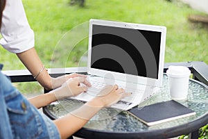 Two woman are seeing laptop on glass table while typing on pad with black note and coffee paper cup in green grass background.