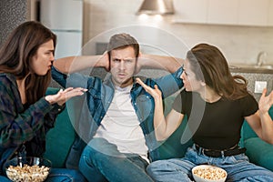 Two women arguing with their friend man sitting together on the sofa, opposite sexes concept