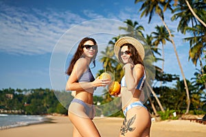 Two women in bikinis hold coconuts on sunny tropical beach with palms. Friends smile, enjoy fresh drinks by sea. Travel