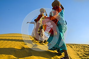 Two women carrying heavy jugs of water on their head and walking on a yellow sand dune