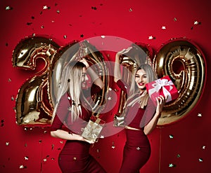 Two women celebrating at new year party happy laughing girls in casual dresses throw gold stars confetti with 2019 balloons