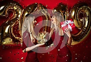 Two women celebrating at new year party happy laughing girls in casual dresses throw gold stars confetti with 2019 balloons
