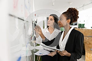 Two women colleagues analyzing data standing next to white board