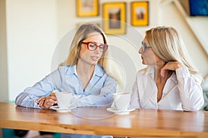 Two women of different generations drink coffee in a cafe and gossip. Girlfriends sit at table chatting and have a