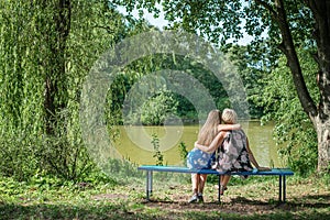 Two women of different generations sitting on a bench near a pond in the summer. Mother and daughter hugging. Grandmother and gran