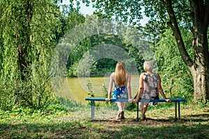 Two women of different generations sitting on a bench near a pond in the summer. Mother and daughter hugging. Grandmother and gran