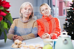 Two women drink tea and eat cakes near the New Year tree.