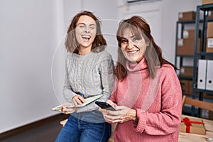 Two women ecommerce business workers writing on notebook using smartphone at office