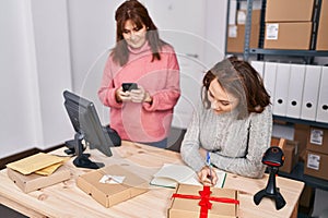 Two women ecommerce business workers writing on notebook using smartphone at office