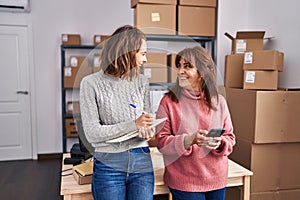 Two women ecommerce business workers writing on notebook using smartphone at office