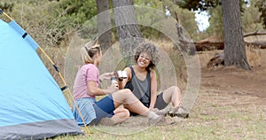 Two women enjoy a camping trip, sharing a laugh over a cup of coffee