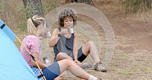 Two women are enjoying a camping trip, sitting by a tent with mugs in hand, with copy space