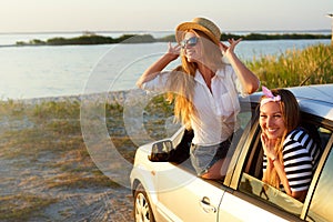 Two women enjoying road trip on a summer day at seaside. Excited young female friends leaning out of car windows and