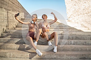 Two women in fashionable sports clothes relaxing after training, taking selfie for social media with mobile phone. Healthy sporty