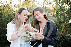 Two women friends eating fresh salads in bowls outdoor together.