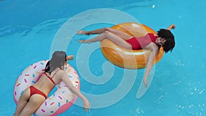 Two women friends foreigners together two people swim in pool at the villa, float on blue water on inflatable lifebuoys