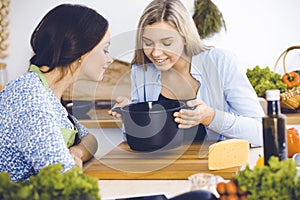 Two women friends looking into the dark pot with a ready meal and taste new recipes while sitting at the kitchen table