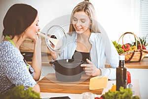 Two women friends looking into the dark pot with a ready meal and taste new recipes while sitting at the kitchen table