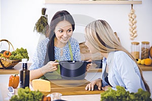 Two women friends looking into the dark pot with a ready meal and taste new recipes while sitting at the kitchen table
