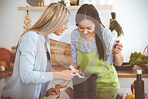 Two women friends looking into the dark pot and taste new recipes for delicious vegetarian soups while standing near the