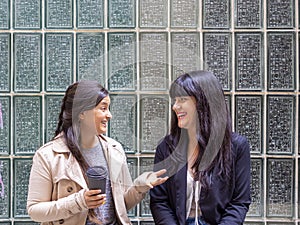 Two women friends sitting outside laughing while drinking a take-away coffee.