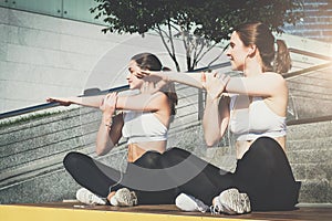 Two women, girls in sportswear doing stretching exercises while listening to music. Workout, couching on city street.
