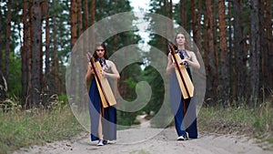 Two women harpists walk at forest and play harps on background of pines.