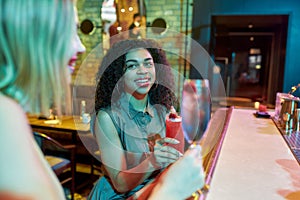 Two women having fun, toasting, drinking cocktails while sitting at the bar counter. Friends spending time at night club