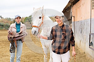 Two women jokey preparing horse for riding in paddock photo