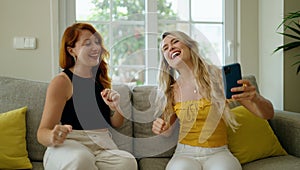 Two women listening to music and dancing sitting on sofa at home