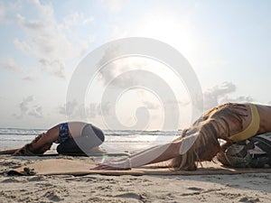 Two women practice child yoga pose at seaside at sunset