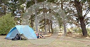 Two women relax beside a blue tent in a wooded campsite with copy space