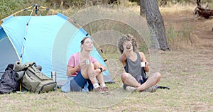 Two women relax by a tent, one holding a mug and the other using binoculars